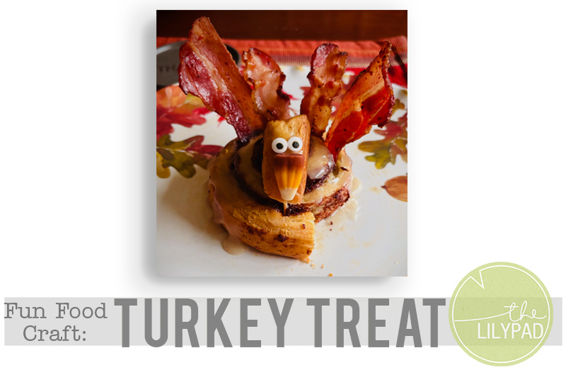 A fun food craft shaped like a turkey made from a cinnamon roll, bacon strips for feathers, and a fruit-and-candy face, displayed on a fall-themed plate with “Fun Food Craft: Turkey Treat” and The Lilypad logo.