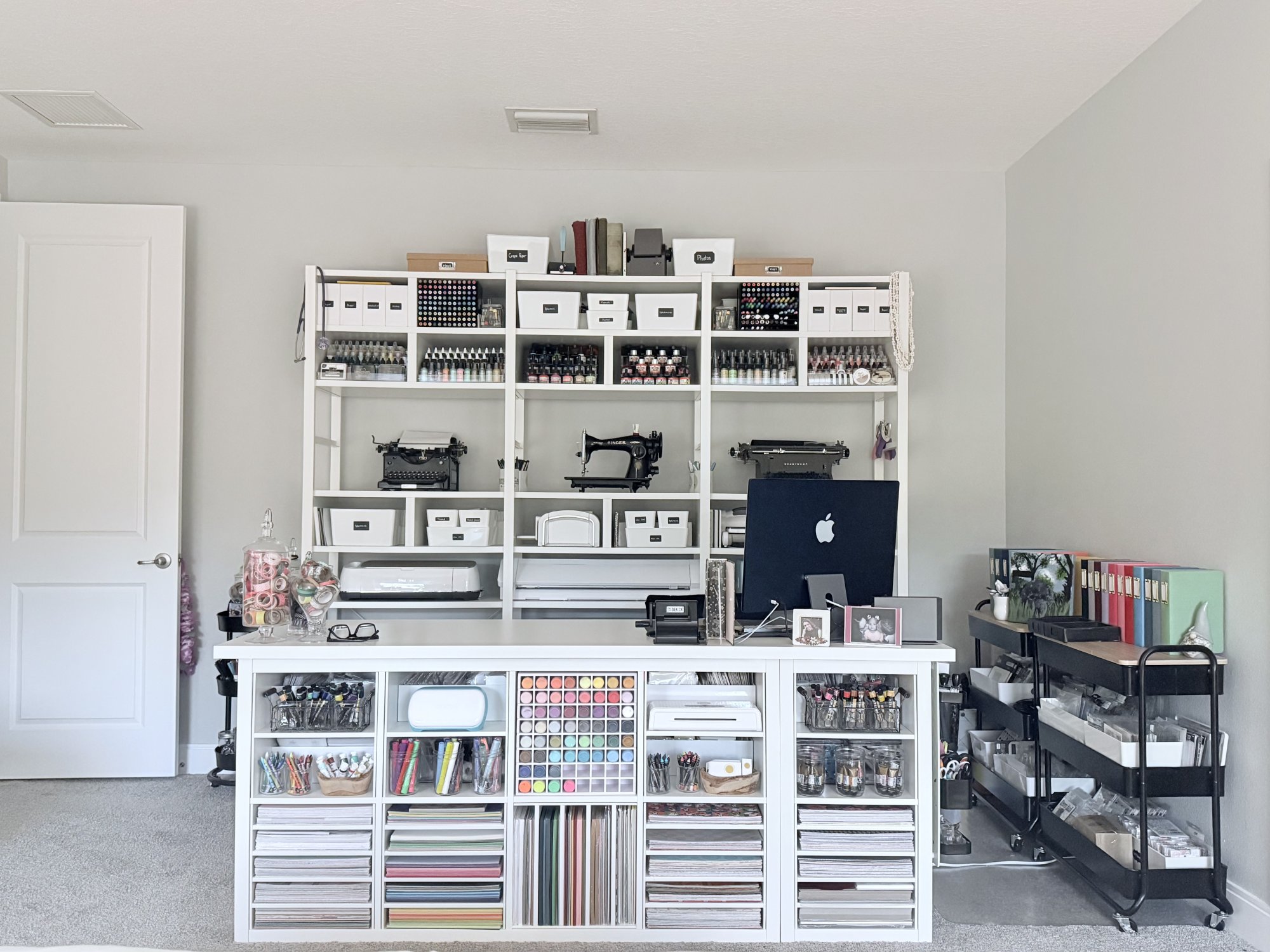 Bright, organized craft room with white shelving filled with labeled storage boxes, rows of ink bottles and markers, vintage typewriters, a sewing machine, paper drawers, and a central worktable with an iMac and neatly arranged supplies.
