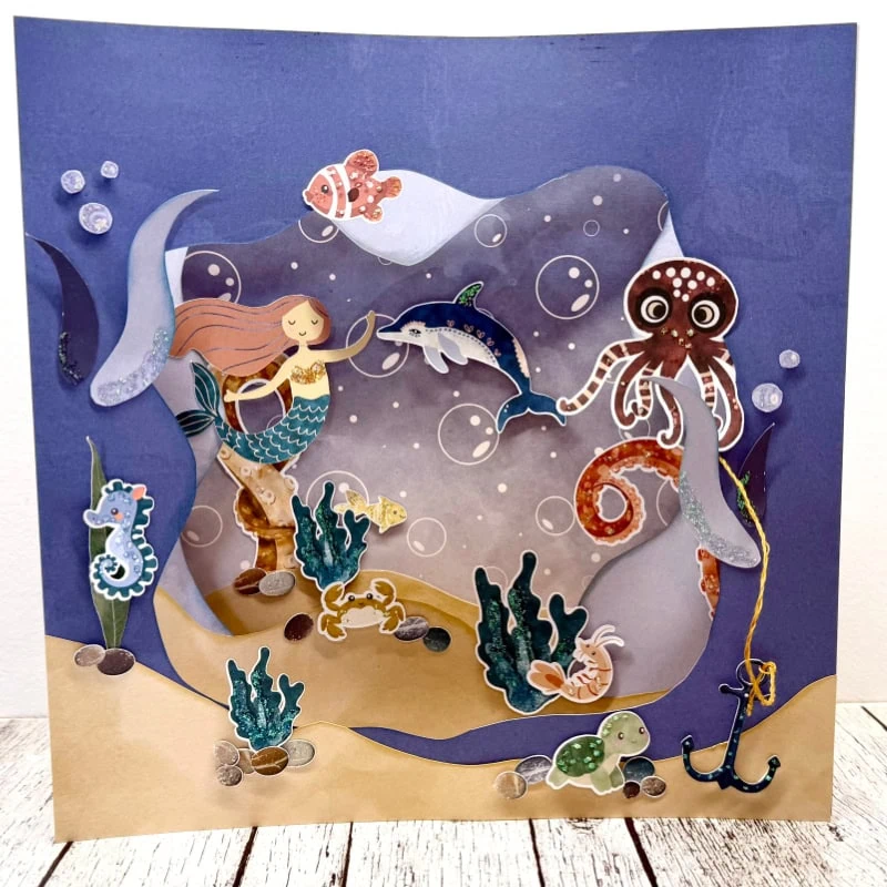 A vibrant layered‑paper diorama of an underwater scene featuring a smiling mermaid with long orange hair and a turquoise tail holding a small dolphin. She is surrounded by colorful sea creatures—including an octopus, seahorse, crab, fish, turtle, and shrimp—along with sea plants, bubbles, rocks, and an anchor resting on the sandy ocean floor.